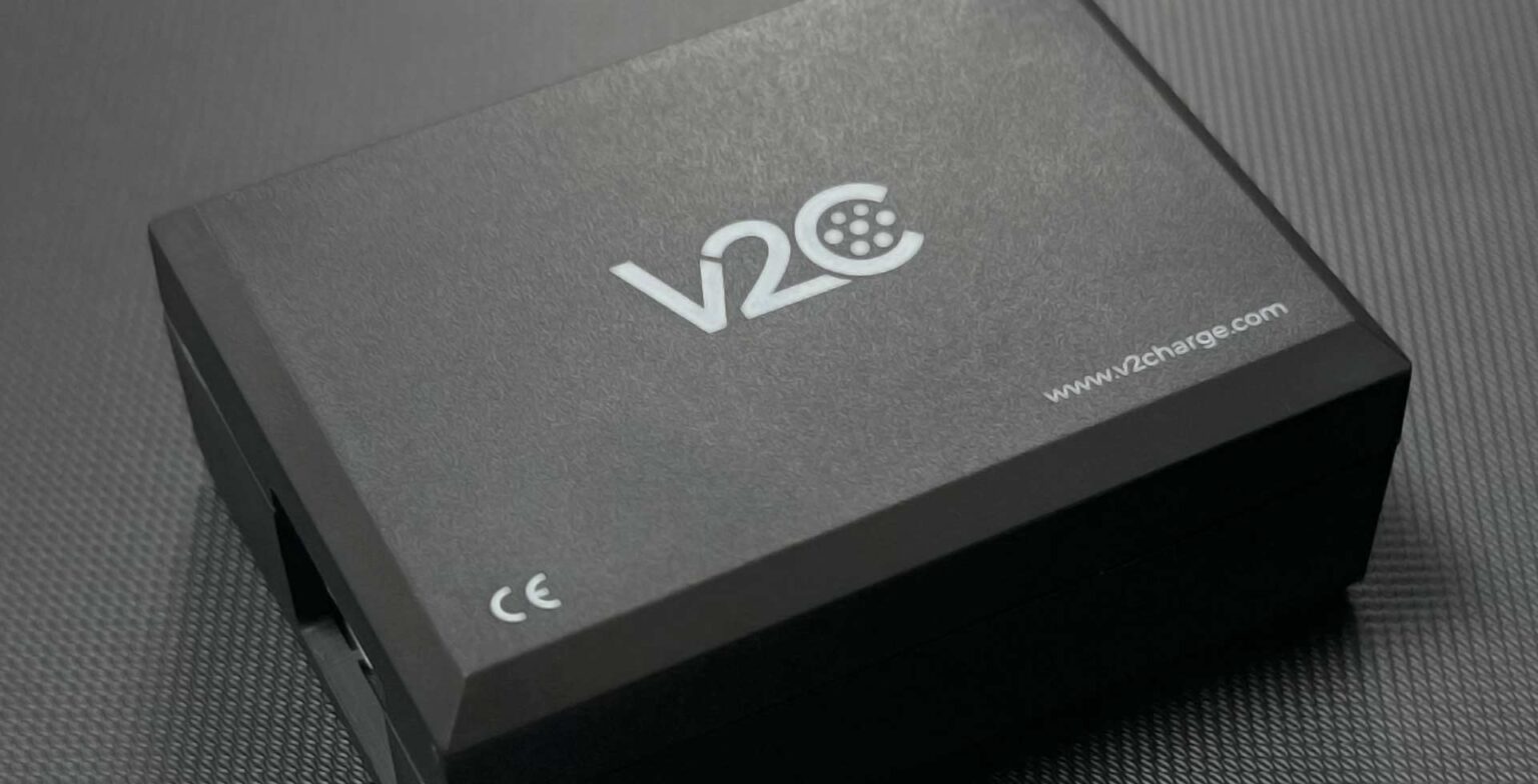 El V2C ControlBox: una caja negra compacta con el logotipo de V2C y el marcado CE, sobre una superficie texturizada gris