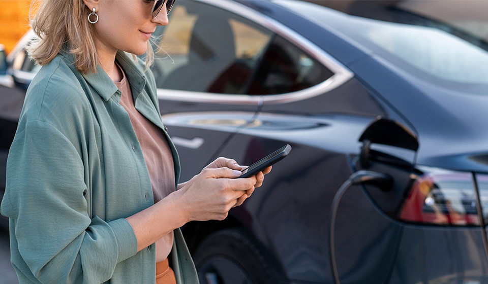 Mujer con el móvil junto a un coche eléctrico enchufado para cargar en las horas más baratas